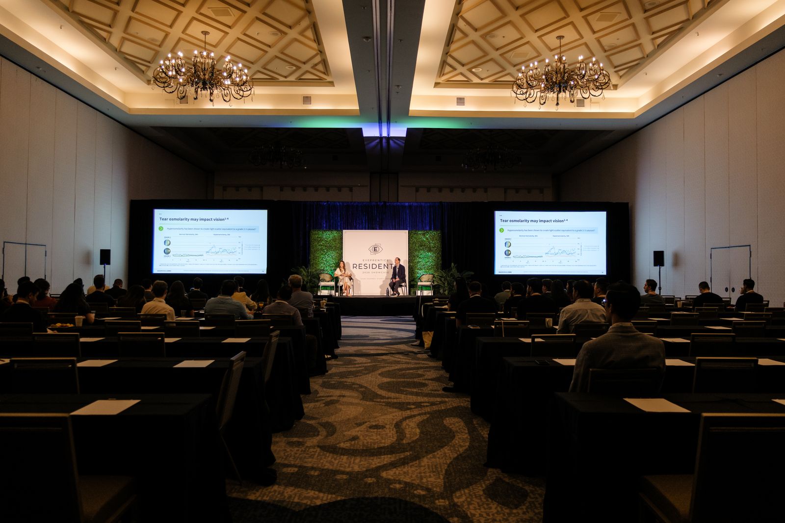 Residents seated in the ballroom watching a main-stage session on dual screens